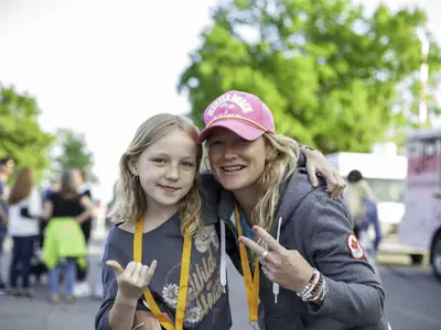 A young girl and mom smiling at the camera giving the peace and rock hand signs.